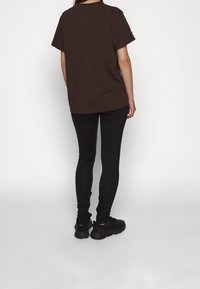 Hnědá polokošile s krátkým rukávem a rovný střih, v kombinaci s černými legínami slim fit a černými teniskami, viděná zezadu.