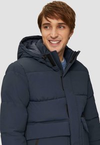 Navyblaue gepolsterte Jacke mit einer Kapuze mit Kordelzug, Reißverschluss und Klettverschlüssen, die große strukturierte Einsätze und eine Fronttasche aufweist.