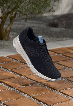 Czarny sportowy sneaker z białą podeszwą i niebieskimi akcentami, unoszący się nad drewnianymi kafelkami na dworze, w tle zielen i kamienne schody.