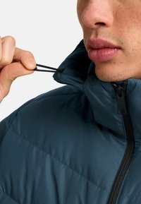 Donker teal gewatteerde jas met een hoge kraag, zwarte rits en een verstelbare trekkoord in de kraag voor een goede pasvorm.