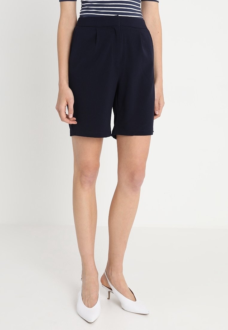 Námořnické šortky na míru s hladkým povrchem, pleteným předním designem a uvolněným střihem. Stylizováno s pruhovaným topem a bílými sandály na podpatku.