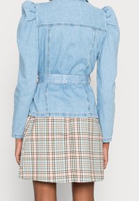 Veste en denim bleu clair avec manches bouffantes et ceinture à la taille, associée à une jupe à carreaux aux couleurs marron, bleu et rouille.
