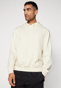 Cream-colored hoodie made of soft fabric, featuring a front pocket and drawstrings. Includes small logo on the chest and ribbed cuffs.