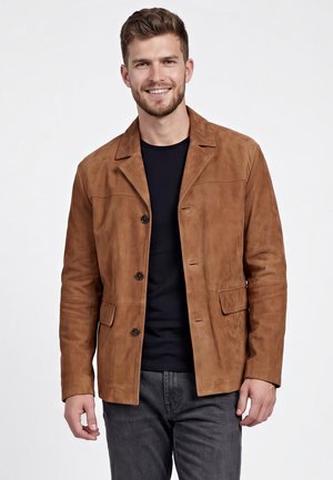 Jonge man glimlacht en draagt een bruine suède jas over een zwart shirt en donkergrijze spijkerbroek tegen een effen witte achtergrond.