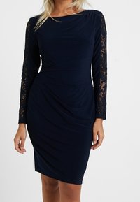 Marinblå klänning med långa spetsärmar, draperad tygdesign och en slät yta. Spetsdetaljerna på ärmarna ger kontrast.