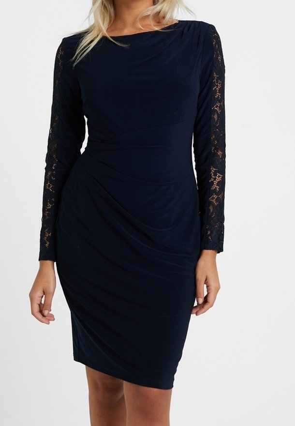 Marinblå klänning med långa spetsärmar, draperad tygdesign och en slät yta. Spetsdetaljerna på ärmarna ger kontrast.