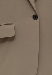 Blazer beige avec un seul bouton noir et une poche à rabat sur le côté gauche.