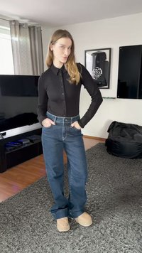 Črna dolgoročna srajca na gumbe, modre jeans hlače z vidnimi šivi in svetli čevlji na vstop, stoji na teksturiranem sivem preprogi.