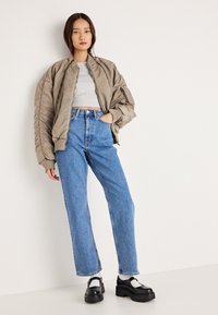 Przesadnie duża taupe bomberka z prążkowanymi mankietami, szary krótki top, niebieskie dżinsy z wysokim stanem i czarne buty na platformie. Gładkie tło.