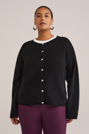 Frau trägt einen schwarzen, geknöpften Cardigan über einem weißen Top und lila Hose, mit großen silbernen Creolen, stehend vor einem einfarbigen Hintergrund.