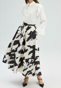Femme portant un chemisier blanc avec des manches à volants et une longue jupe imprimée abstraite noire et blanche avec des talons noirs pointus.