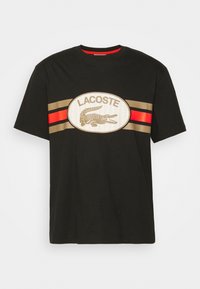 Černé bavlněné tričko s velkým oválným logem Lacoste a grafikou krokodýla, zvýrazněné zlatými a červenými horizontálními pruhy přes hruď.