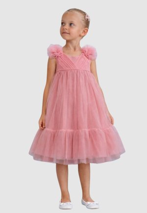 Ragazza giovane in piedi, indossa un vestito rosa in tulle con dettagli a spalla gonfia e scarpe da balletto bianche, guarda leggermente di lato.