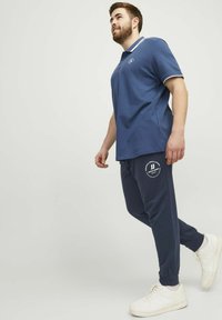 Marinblå polotröja med vita krageaccent, i kombination med marinblå sweatpants med en vit cirkulär logotyp, kompletterat med vita sneakers.