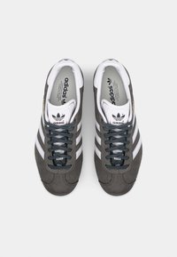 Grå mockaskor med tre vita ränder, mörk snörning och en vit tunga med Adidas-logotyp. Slät innerfoder. Klassisk design.