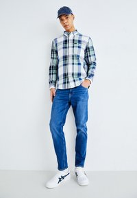 Koszulka w kratę w kolorze zielonym, niebieskim i białym; niebieskie dżinsy; białe sneakersy z granatowymi akcentami; czapka dżinsowa. Prosty, casualowy strój.
