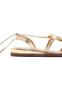 Sandalias metálicas doradas con tiras finas y un acento decorativo de perla en la parte frontal. Suela plana con un detalle en el tacón de tono dorado.