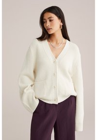 Cardigan lavorato a maglia di colore crema con scollo a V, tre bottoni e tasche frontali, abbinato a pantaloni larghi viola.