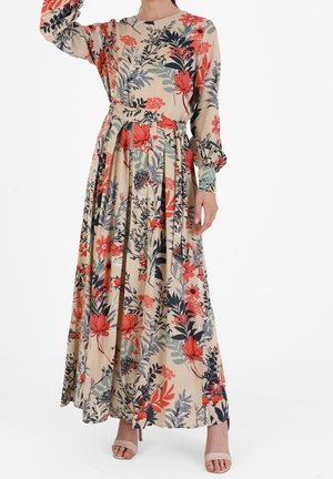 Kvinde iført en lang beige blomstret kjole med røde, sorte og grønne blade og blomster, kombineret med beige åbne hæle.