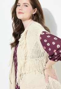 Strickweste in Creme mit Fransenakzenten, über einer tiefvioletten Bluse mit blühenden Ärmeln getragen. Texturierte Maschen sind sichtbar.