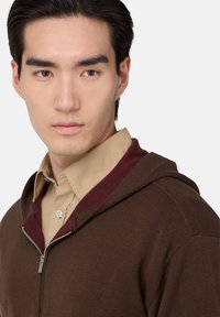 Hnedá mikina na zips s bordovým akcentom na kapucni, nosená cez béžovú košeľu s golierom. Tkanina vyzerá mäkko s hladkým povrchom.