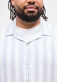 Άνδρας με γενειάδα και κοτσιδάκια που φοράει λευκό πουκάμισο με ανοιχτό μπλε κάθετες ρίγες και λευκά κουμπιά πάνω από λευκό μπλουζάκι.