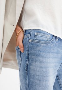 Vybledlé modré džíny s úzkým střihem, se přední kapsou, stříbrnými knoflíky a kontrastním švem. Nosené s bílým topem.