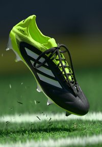 Black and neon green football boot with three white stripes, textured upper, laces, and molded studs, suspended above grass with flying debris.