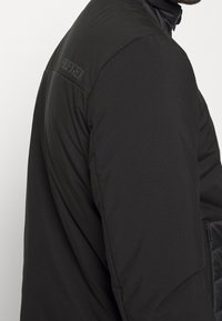 Black jacket featuring a smooth texture, high collar, and embroidered logo on the shoulder. Zip pocket on the lower side adds functionality.