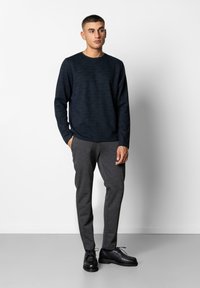 Navyfarbendes, strukturiertes Langarmshirt kombiniert mit grau geschnittenen Hosen und schwarzen Schnürschuhen, vor einem schlichten weißen Hintergrund.