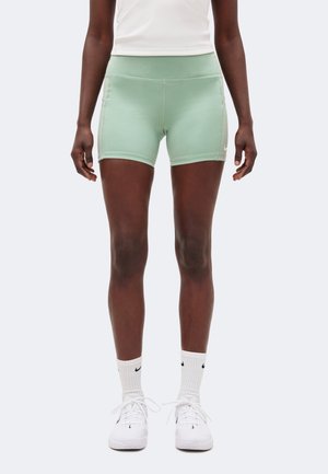 Kvinde iført lysegrønne tætsiddende atletikshorts, hvide sokker og hvide sneakers, stående mod en ensfarvet baggrund.