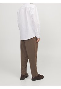 Camisa blanca de manga larga con puños abotonados, combinada con pantalones marrones holgados. El conjunto se completa con zapatos oscuros.