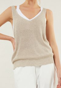 Haut sans manches beige en tricot avec un décolleté en V, doté d'une coupe ample et d'un ourlet côtelé. Superposé sur un débardeur blanc. Texture lisse.