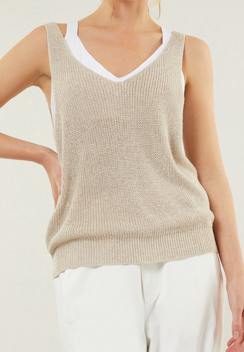 Haut sans manches beige en tricot avec un décolleté en V, doté d'une coupe ample et d'un ourlet côtelé. Superposé sur un débardeur blanc. Texture lisse.