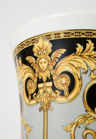 Xícara de cerâmica com padrões barrocos intrincados em dourado e preto, apresentando um motivo de rosto proeminente e detalhes florais, acabamento liso e brilhante.