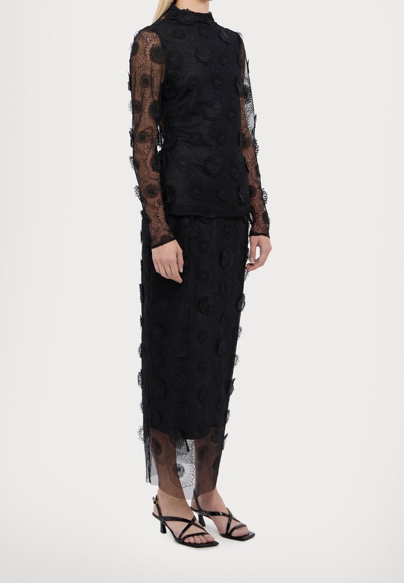 Femme portant un haut en dentelle noire à manches longues et une jupe midi assortie avec des applications florales, ainsi que des sandales à talons noires à brides.