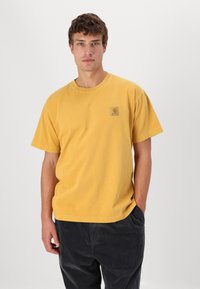 T-shirt di cotone giallo con maniche corte, vestibilità rilassata e un patch quadrato con logo sul lato sinistro del petto. Texture liscia. Stile casual.