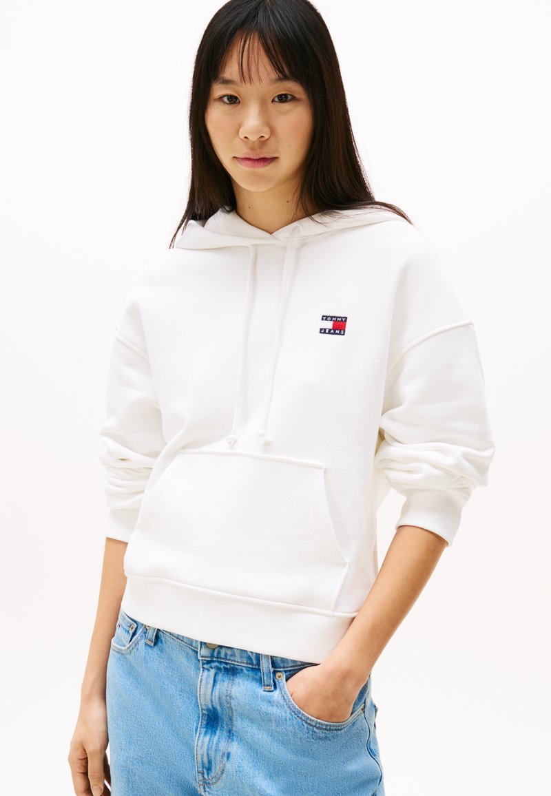 Hvit hettegenser med frontlomme og snøring i hetten. Har et lite rødt og blått logo-patch på brystet. Blå jeans med en falmet utseende.