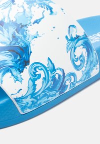 Niebieska klapka na ślizg z białym paskiem ozdobionym niebieskimi falami oraz fakturowaną niebieską podeszwą. Zaprojektowana do codziennego noszenia z miękkim wykończeniem.