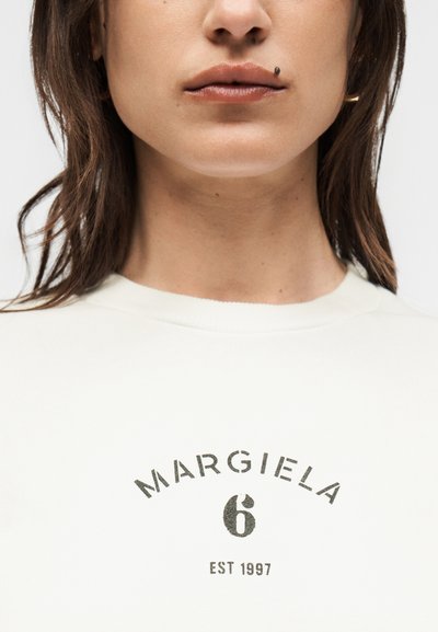 Κοντινό πλάνο ενός ατόμου που φορά λευκό πουκάμισο με μαύρη εκτύπωση "MARGIELA 6 EST 1997", με καστανά μαλλιά και χρυσούς κρίκους στα αυτιά.
