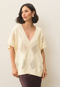 Ohlapen pulover kremne barve z V-izrezom, okrašen z vzorci čipke v obliki diamantov in rebrastimi manšetami, kombiniran s temno krilo.