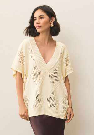Ohlapen pulover kremne barve z V-izrezom, okrašen z vzorci čipke v obliki diamantov in rebrastimi manšetami, kombiniran s temno krilo.