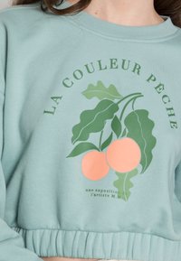 Sudadera corta en verde menta con gráficos de melocotón y hojas, y el texto "LA COULEUR PÊCHE", con puños acanalados en el dobladillo.