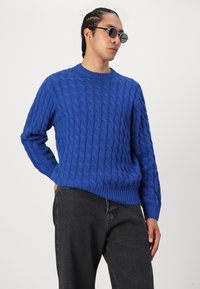 Blue cable-knit sweater with round neckline and ribbed cuffs, paired with dark jeans. Model wears round sunglasses.