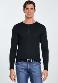 Vyras dėvi juodą ilgomis rankovėmis henley marškinėlį su trimis sagomis ir mėlynas džinsines kelnes, stovi priešais šviesų paprastą foną.