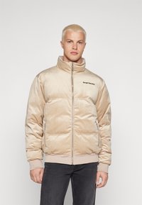 Giacca imbottita beige con collo alto, chiusura a zip e logo sul lato sinistro; realizzata in materiale morbido e testurizzato; polsini e orlo a coste.