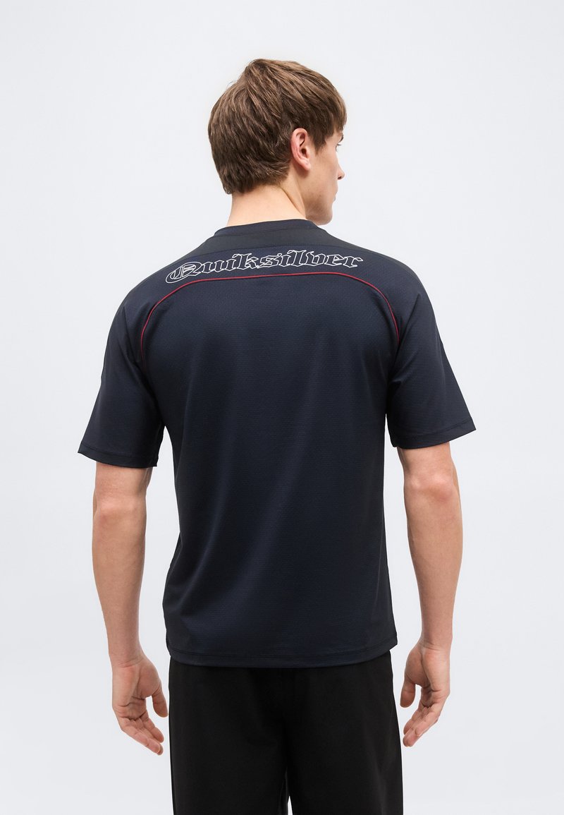 Άνδρας που φαίνεται από πίσω, φοράει μαύρο t-shirt Quiksilver με κοντά μανίκια και κόκκινη ραφή κατά μήκος των ραφών στους ώμους.