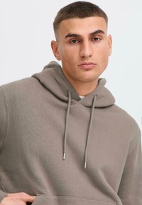 Young man with short, styled hair wearing a light brown hoodie, looking directly at the camera with a neutral expression against a plain backdrop.