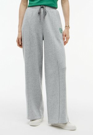 Donna che indossa pantaloni della tuta grigio chiaro a gamba larga con coulisse in vita e sneakers bianche, in piedi su sfondo bianco.