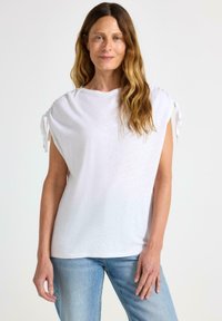 Hvid kortærmet top med samlede skuldre, lavet af et blødt stof. Har bundet detaljer på ærmerne og en afslappet pasform. Bæres med blå jeans.